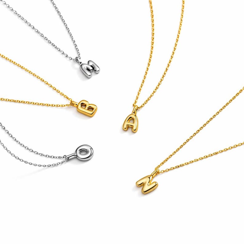 May include: A collection of gold and silver-toned initial necklaces. The necklaces feature the letters A, B, M, O, and N. Each letter is a pendant on a delicate chain. The pendants have a rounded, bubble-like design.