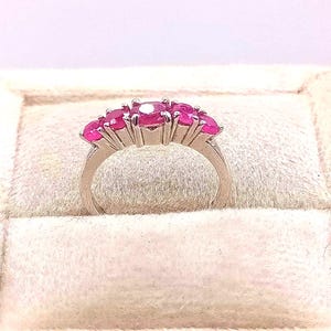 May include: A silver ring featuring a row of vibrant pink gemstones. The ring's band curves gracefully, and the gemstones are set closely together, creating a continuous line of color. The ring is displayed on a soft, light-colored surface.