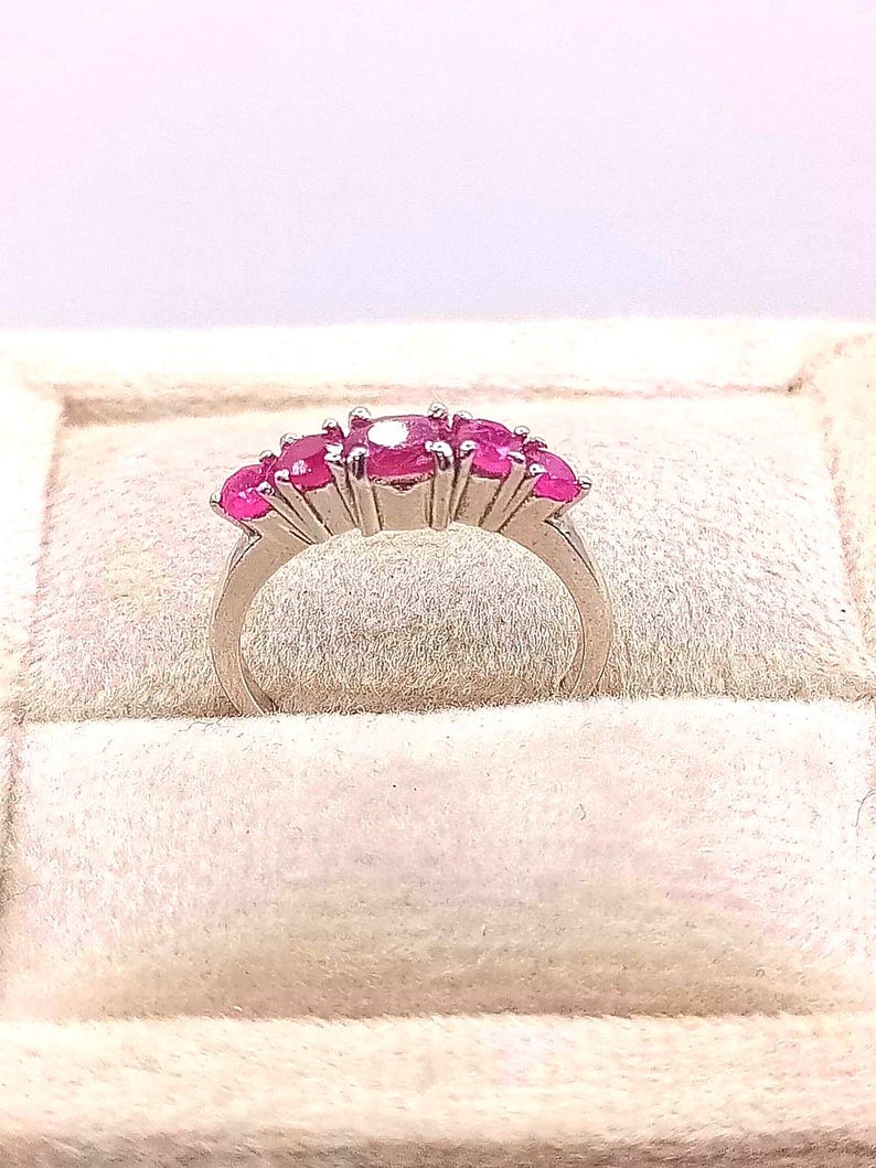 May include: A silver ring featuring a row of vibrant pink gemstones. The ring's band curves gracefully, and the gemstones are set closely together, creating a continuous line of color. The ring is displayed on a soft, light-colored surface.