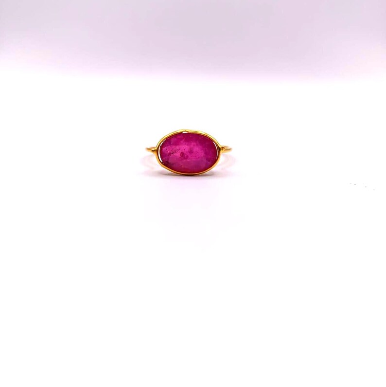 May include: A gold ring featuring a large, oval-shaped, vibrant pink gemstone. The gemstone is the focal point, set in a simple, elegant gold band. The ring is displayed on a white background.