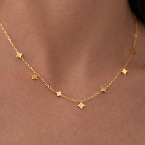 Pode incluir: Um delicado colar dourado com pequenos pingentes em forma de diamante. O colar é suspenso por uma corrente fina, criando um design minimalista e elegante. O colar é mostrado em um fundo de tom de pele.