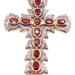 May include: A detailed, ornate cross pendant featuring oval-cut, deep red gemstones set in gold bezels. The cross is embellished with silver-toned metal and small, sparkling accents, creating a luxurious and eye-catching design.
