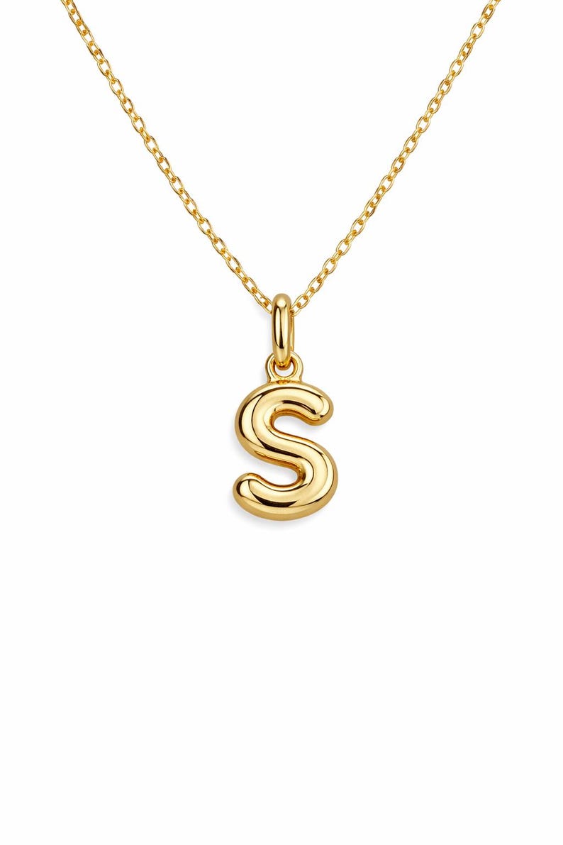 May include: A gold-colored necklace featuring a pendant in the shape of the letter "S". The pendant is a bold, rounded design, suspended from a delicate chain. The necklace is a simple yet elegant piece of jewelry.
