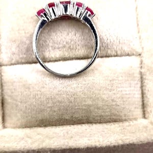 May include: A silver ring featuring five vibrant red gemstones. The gemstones are set in a row across the top of the ring, creating a continuous line of color. The ring's band is smooth and polished, reflecting light.