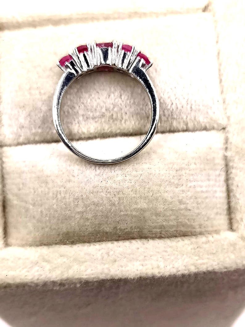 May include: A silver ring featuring five vibrant red gemstones. The gemstones are set in a row across the top of the ring, creating a continuous line of color. The ring's band is smooth and polished, reflecting light.