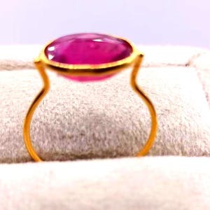 May include: A gold ring featuring a large, oval-shaped, vibrant pink gemstone. The gemstone is set in a gold bezel, and the ring's band has a unique, open design. The ring is displayed on a soft, textured surface.