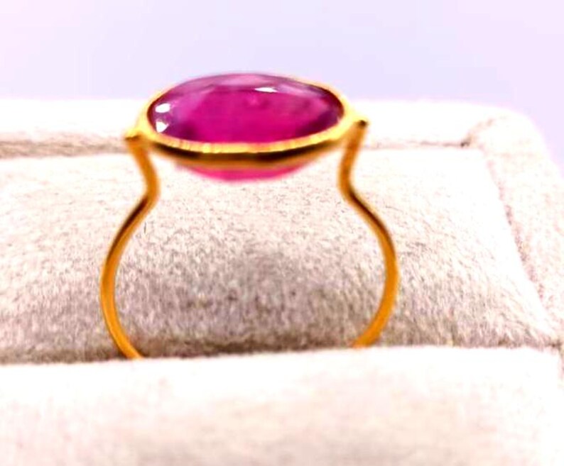 May include: A gold ring featuring a large, oval-shaped, vibrant pink gemstone. The gemstone is set in a gold bezel, and the ring's band has a unique, open design. The ring is displayed on a soft, textured surface.