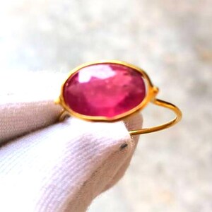 May include: A gold ring featuring a large, oval-shaped, pink gemstone. The gemstone is set in a gold bezel, and the ring has a simple, thin gold band. The ring is displayed against a white background.
