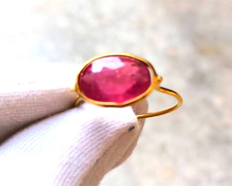 May include: A gold ring featuring a large, oval-shaped, pink gemstone. The gemstone is set in a gold bezel, and the ring has a simple, thin gold band. The ring is displayed against a white background.