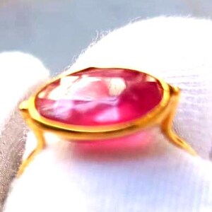 May include: A gold ring featuring a large, oval-cut, pink gemstone. The gemstone is set in a gold bezel setting with delicate, curved prongs. The ring is displayed against a white background, highlighting the vibrant color of the stone.