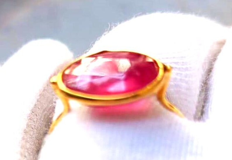 May include: A gold ring featuring a large, oval-cut, pink gemstone. The gemstone is set in a gold bezel setting with delicate, curved prongs. The ring is displayed against a white background, highlighting the vibrant color of the stone.