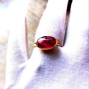 May include: A gold ring featuring a large, oval-cut ruby gemstone. The ruby is a deep red color and is set in a simple gold band. The ring is displayed on a white surface.
