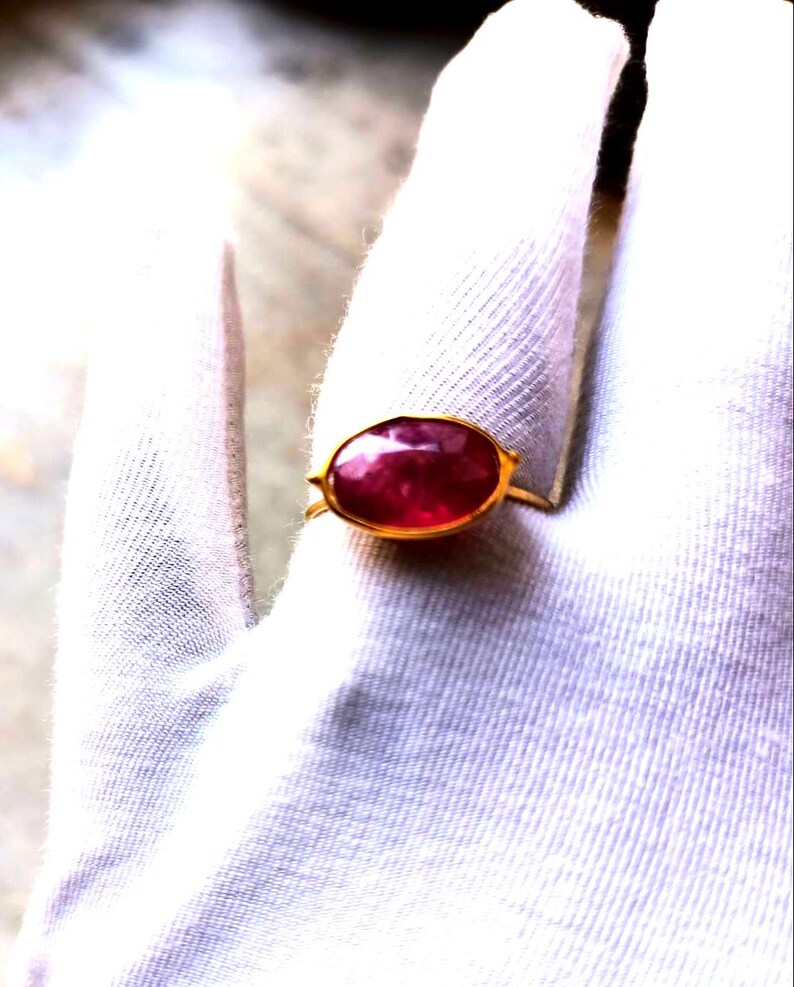 May include: A gold ring featuring a large, oval-cut ruby gemstone. The ruby is a deep red color and is set in a simple gold band. The ring is displayed on a white surface.