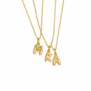 May include: Three gold-colored necklaces with pendant charms spelling out the letters "MKA". Each necklace has a delicate chain and a rounded, bubble-style letter charm. The background is plain white.