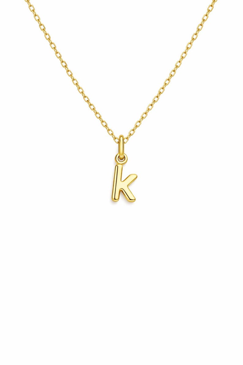 May include: A gold-colored necklace featuring a small pendant in the shape of the letter "K". The pendant and chain are both a bright, shiny gold. The chain is delicate and appears to be made of small, connected links.