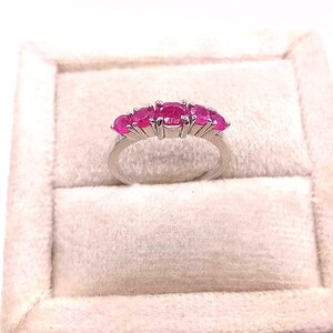May include: A silver ring featuring five oval-cut, vibrant pink gemstones. The gemstones are set in a row across the top of the ring. The ring is displayed on a light-colored, textured surface, likely a jewelry box.