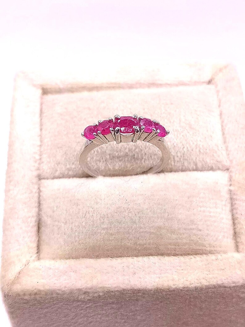 May include: A silver ring featuring five oval-cut, vibrant pink gemstones. The gemstones are set in a row across the top of the ring. The ring is displayed on a light-colored, textured surface, likely a jewelry box.