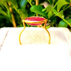 May include: A gold-colored ring with a large, oval, red gemstone. The gemstone is set in a gold bezel and supported by delicate, curved gold prongs. The ring is displayed against a white background with green foliage in the background.