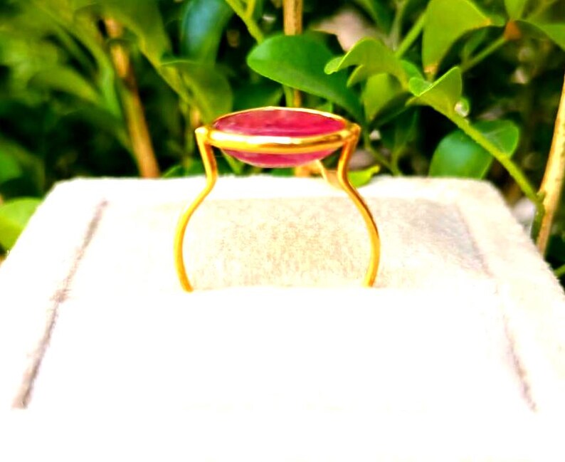 May include: A gold-colored ring with a large, oval, red gemstone. The gemstone is set in a gold bezel and supported by delicate, curved gold prongs. The ring is displayed against a white background with green foliage in the background.