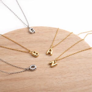 May include: A collection of gold and silver initial necklaces displayed on a wooden surface. The necklaces feature the letters G, B, A, O, and Z. The chains are delicate, and the pendants are in a bold, rounded font.