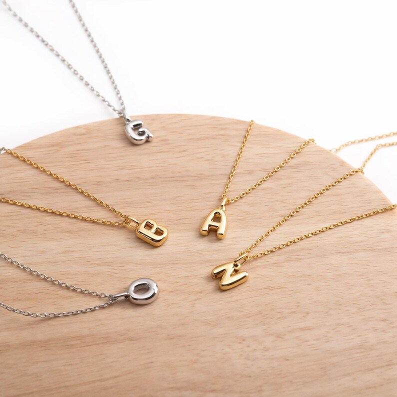 May include: A collection of gold and silver initial necklaces displayed on a wooden surface. The necklaces feature the letters G, B, A, O, and Z. The chains are delicate, and the pendants are in a bold, rounded font.