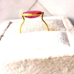 May include: A gold ring featuring a large, oval-shaped, pink gemstone. The gemstone is set in a gold bezel, and the ring has a delicate, curved band. The ring is displayed on a white, textured surface.