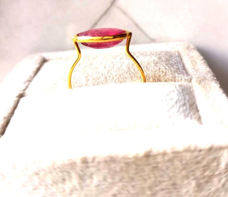 May include: A gold ring featuring a large, oval-shaped, pink gemstone. The gemstone is set in a gold bezel, and the ring has a delicate, curved band. The ring is displayed on a white, textured surface.