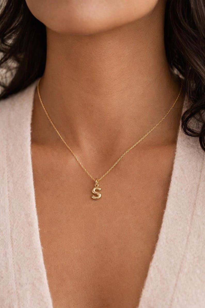 May include: A gold-colored necklace with a pendant in the shape of the letter "S". The pendant hangs from a delicate chain. The necklace is worn around the neck.