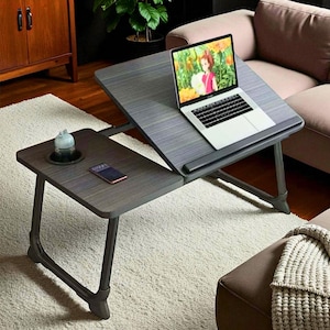 May include: A grey, adjustable laptop desk with a separate side table. A laptop displaying a colourful image sits on the main surface. A cup and a smartphone rest on the side table. The desk has dark grey legs and is on a white rug.