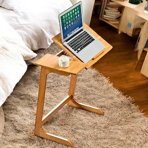 Pode incluir: Uma mesa de madeira para laptop com uma superfície inclinável e uma pequena mesa lateral. Uma caneca branca está na mesa lateral. Um laptop prateado está aberto na mesa. A mesa está em um tapete marrom.