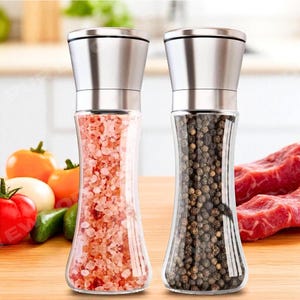 May include: Two glass salt and pepper grinders with stainless steel tops. One filled with pink Himalayan salt, the other with black peppercorns. The grinders are on a wooden surface with vegetables and meat in the background.