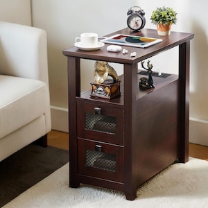 Puede incluir: Una mesa auxiliar de madera marrón oscuro con dos cajones y un estante. El estante superior contiene una tableta, una taza y un platillo, y un pequeño despertador. Un gramófono en miniatura se encuentra en el estante de abajo. La mesa está sobre una alfombra blanca.
