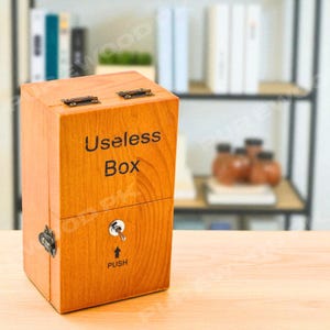 Puede incluir: Caja de madera clara con la inscripción "Useless Box". Tiene una tapa con bisagras y cierres metálicos, y un interruptor con la palabra "PUSH". El diseño es simple y rectangular, con vetas de madera.