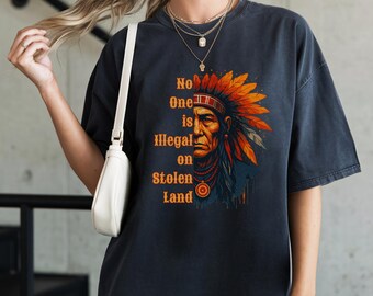 Może przedstawiać: Ciemnoszara koszulka z portretem Indianina w pióropuszu w kolorze pomarańczowym i żółtym. Tekst "No One is Illegal on Stolen Land" jest wydrukowany na pomarańczowo. Koszulka ma wygląd vintage.