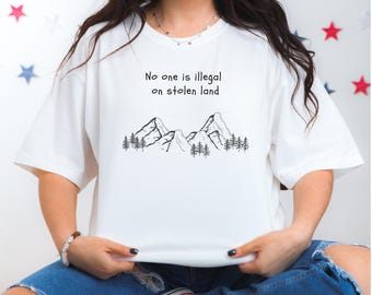 Może przedstawiać: Biała koszulka z napisem "No one is illegal on stolen land" nad czarno-białą grafiką gór i sosen. Koszulka ma luźny, obszerny krój.