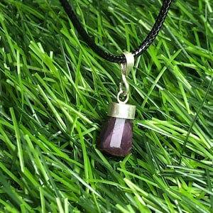 Könnte beinhalten: Eine Anhängerkette mit einem dunkelvioletten Edelstein und einer silberfarbenen Kappe. Der Edelstein hängt an einer schwarzen Kordel. Die Kette wird vor einem Hintergrund aus grünem Gras präsentiert.