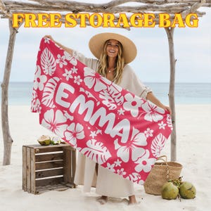 May include: A red beach towel featuring white floral designs and the name "EMMA" in white lettering. The towel is displayed on a beach, with the words "FREE STORAGE BAG" in yellow at the top.