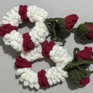 May include: Crocheted garland with a festive design. The garland features alternating sections of white and burgundy, with a ruffled texture. Crochet roses in burgundy and green are attached to the garland.