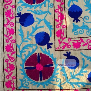 May include: Close-up of a beige textile with intricate embroidery. The design features large, stylized flowers in shades of fuchsia and deep purple, surrounded by light blue foliage and dark blue pomegranate shapes. A border of fuchsia flowers frames the design.