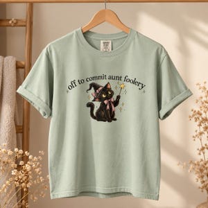 May include: A light sage green short-sleeve t-shirt featuring a graphic of a black kitten wearing a witch hat and a pink bow, holding a star wand. The text above the kitten reads, off to commit aunt foolery, in a dark brown serif font.