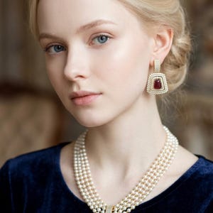 May include: A multi-strand pearl necklace featuring a large, rectangular pendant with a deep red centre stone surrounded by clear crystals, paired with matching rectangular drop earrings that feature the same red stone and crystal design.