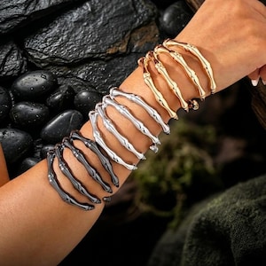 Könnte beinhalten: Ein Set aus drei Metallarmbändern in Form von Skelettfingern mit goldfarbenem, silberfarbenem und schwarzem Finish. Die Armbänder sind am Arm gestapelt und der Text Aussies Aura ist am unteren Rand des Bildes aufgedruckt.