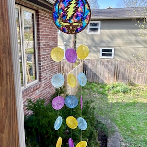 May include: A colorful wind chime featuring a round wooden top with a Steal Your Face design. The design is a skull with a lightning bolt. Below, iridescent shell-like discs in yellow, purple, and blue hang from strings, creating a decorative outdoor accent.