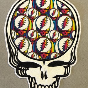 May include: A white skull-shaped patch with a black outline and teeth. The skull's interior is filled with smaller skulls, each featuring a rainbow-colored lightning bolt design on a white background. The patch has a soft, textured appearance.