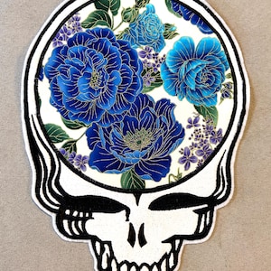 May include: A white patch in the shape of a skull with a black outline and details. The skull's head contains a floral design with large blue flowers, smaller purple flowers, and green leaves. The patch has a black and white striped border.