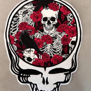 May include: A white and black skull-shaped patch with a black outline. The interior features a skull, red roses, a raven, and a skeleton design. The patch has a white border with black stripes.