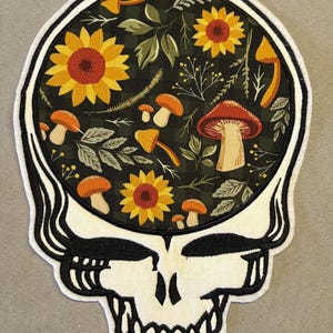 May include: A skull-shaped patch with a black and white outline. The skull's interior features a floral design with sunflowers, mushrooms, and foliage on a dark green checkered background. The patch has a white border.