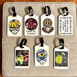 May include: Eight white luggage tags with black straps, each featuring a unique design. The designs include a skull with a palm tree, a figure with the text "Keep On Truckin'," and a skull with an eye.
