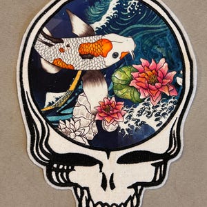 May include: A white and black skull-shaped patch with a detailed illustration inside. The illustration features a koi fish, lotus flowers, and ocean waves. The koi fish has orange and white scales, and the flowers are pink and green.