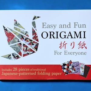 以下が含まれることがあります： 「Easy and Fun ORIGAMI」と日本語の文字が書かれた白い箱。左側にはカラフルな折り鶴があります。箱には、伝統的な日本の模様の折り紙が28枚入っています。